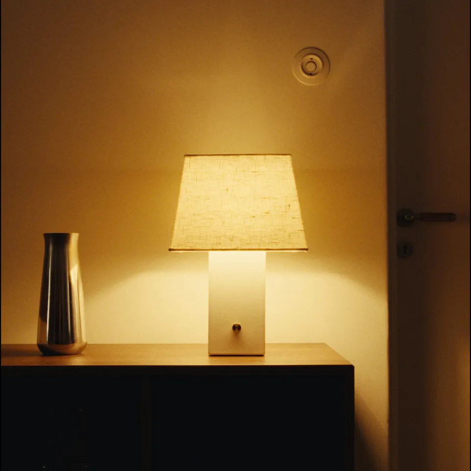 A modern table lamp, HALGATT, with beige linen shade and white ceramic base. Its turned on, casting warm light.