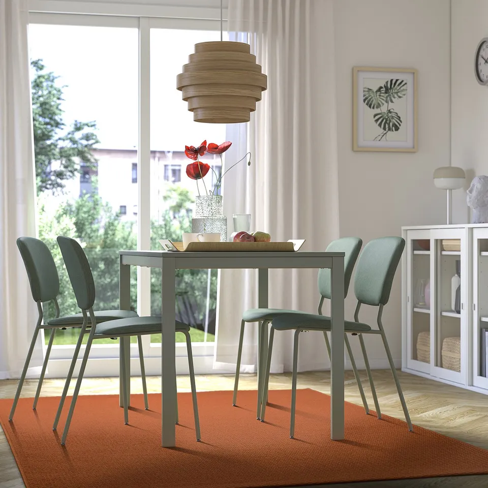 Modern dining room with glass table, green chairs, wooden pendant light, floral centrepiece, orange rug, large window.