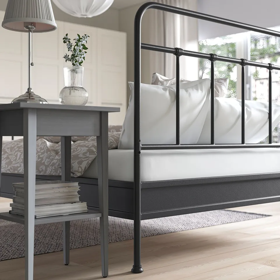 Modern bed with high headboard, pillows. Beside table with lamp, books, flowers. Large windows with natural light.
