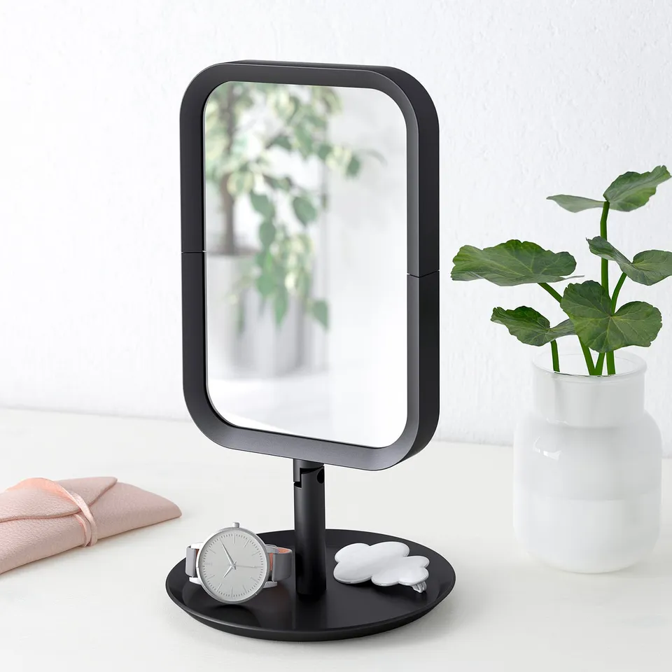 A black vanity mirror with magnified side, watch, towel, and plant on its base.