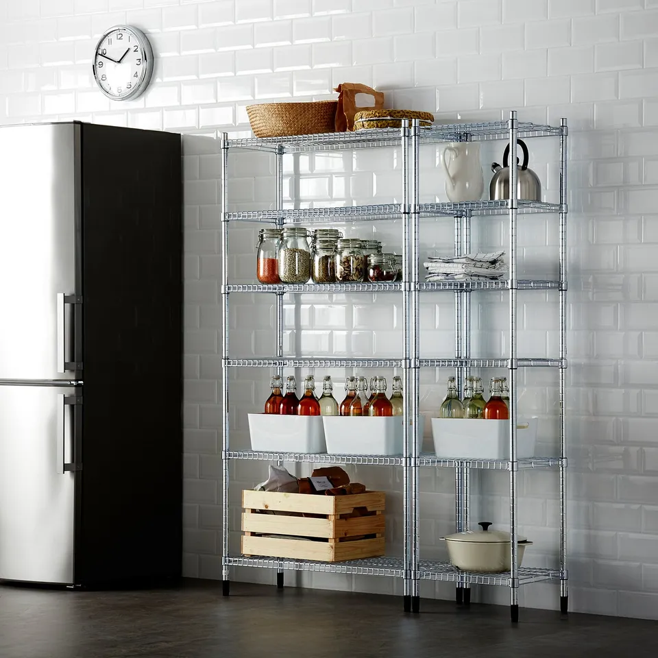 Metal shelving unit against white tiled wall, holding jars, bottles, baskets, and kitchen items, next to tall fridge.