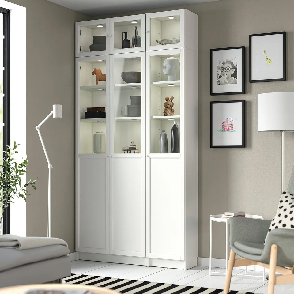 White BILLY bookcase with glass doors, displaying decor. Set in grey living room with sofa, chair, and wall art.
