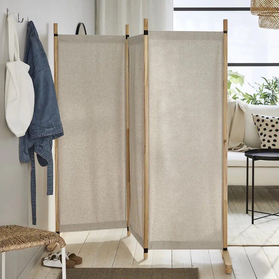 The GLAMBERGET beige room divider with light wooden frames, dividing a cosy entryway with hanging jackets and a seating area.