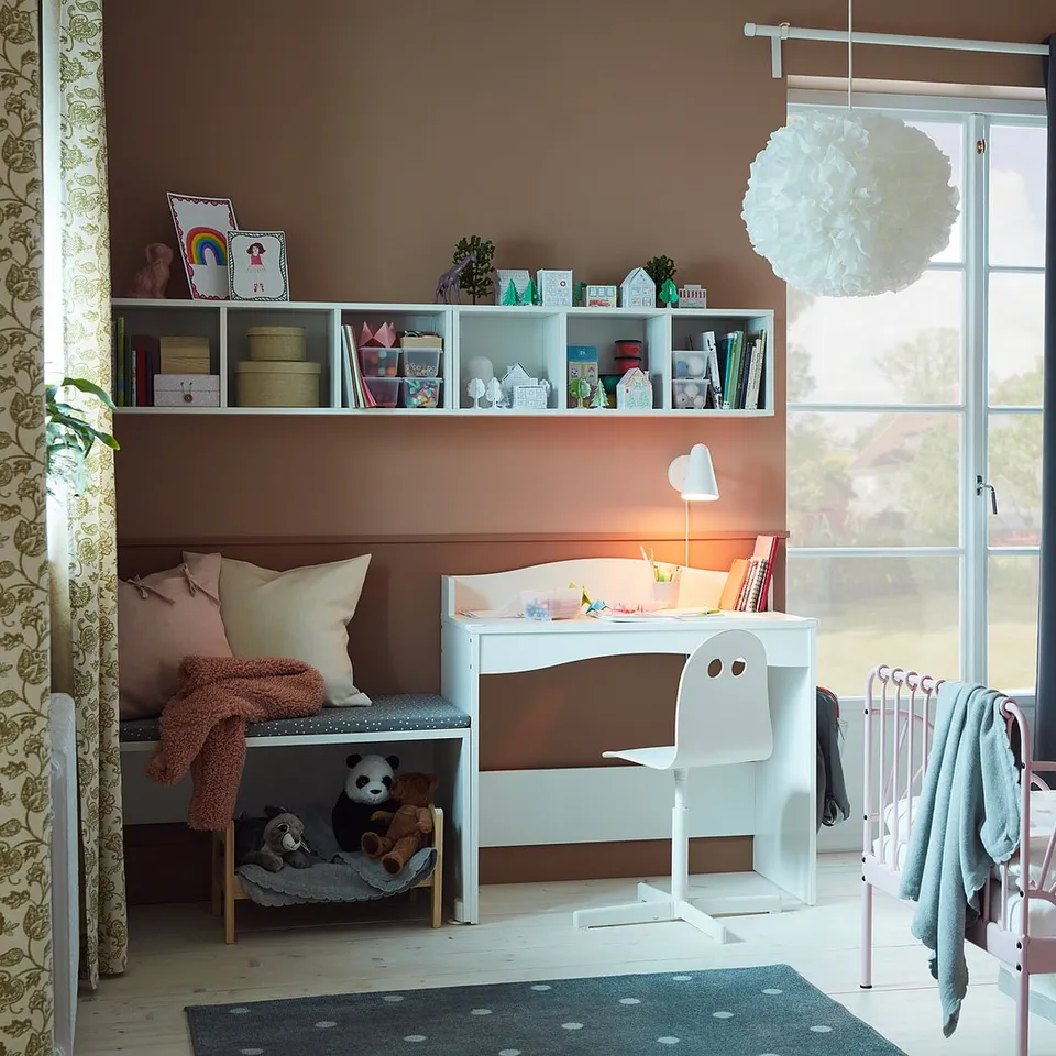 Bright childrens room with white SMÅGÖRA desk, chair, and bed, peach walls, shelves with books and toys, cosy pillows, and large window.