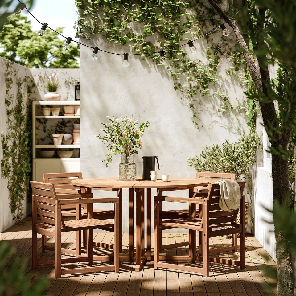 Outdoor dining set with 4 wooden chairs and table, under string lights and a white wall with greenery.