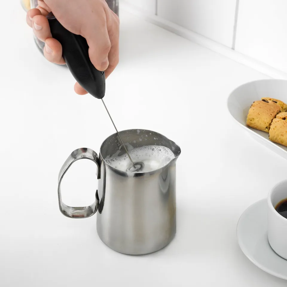 A hand uses an electric frother to create foam in a metal pitcher, enhancing coffee with a creamy texture.