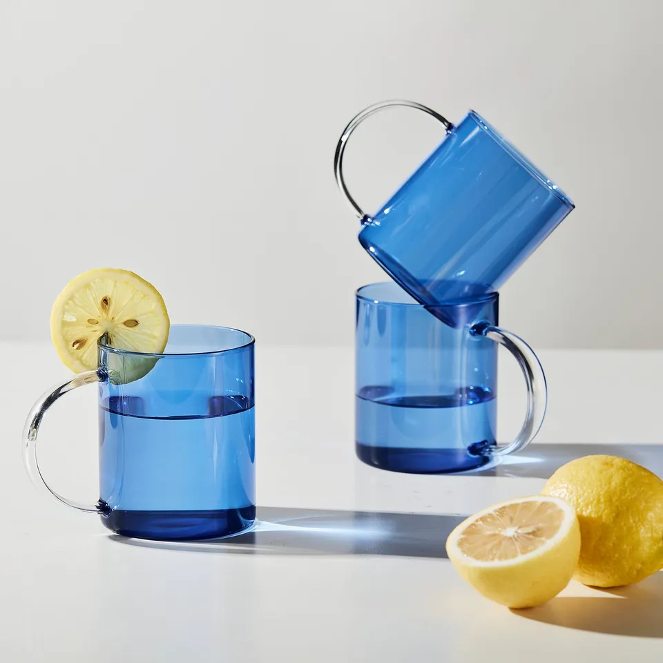 A set of blue glass mugs; one upright with lemon slice, another tilted, showcasing their heat-resistant, multi-temperature use.