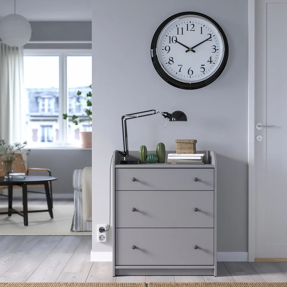 Grey BRAVUR wall clock above drawer unit in bright room.