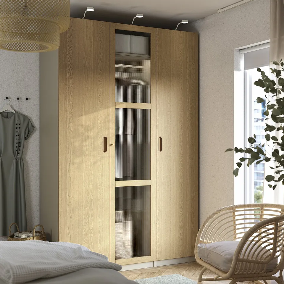Modern bedroom with large PAX wardrobe in light wood. Opposite, a wicker chair beside a large window.