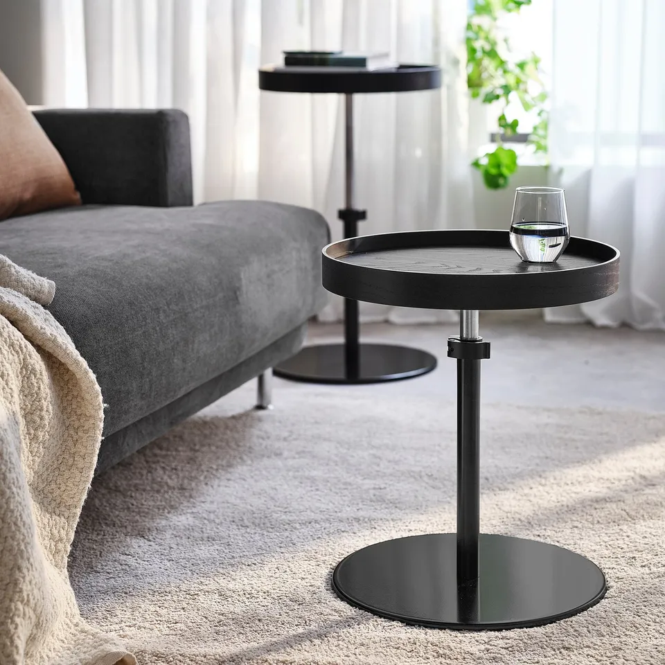 A modern living room a grey sofa with a black side table next to it. The adjustable table holds a wine glass and has a black base and top.