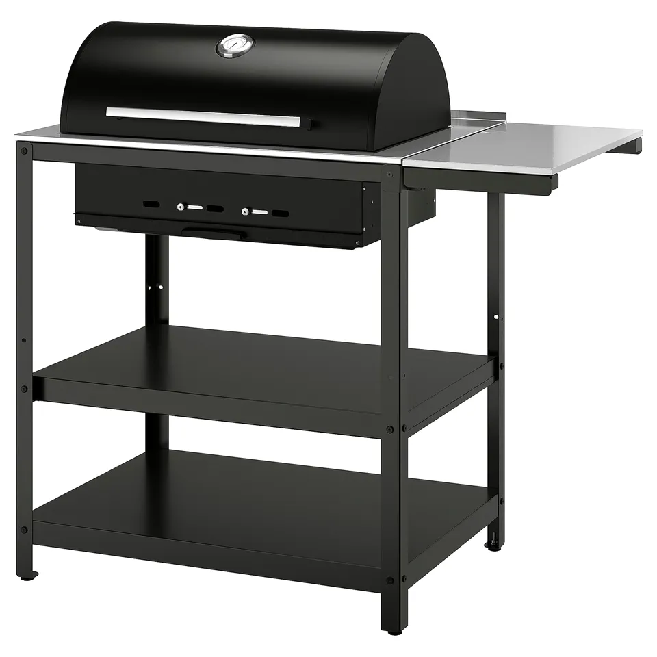 Black GRILLSKÄR charcoal grill with thermometer, stainless steel work surfaces, and three shelves. Insulated handles and side table attachment.