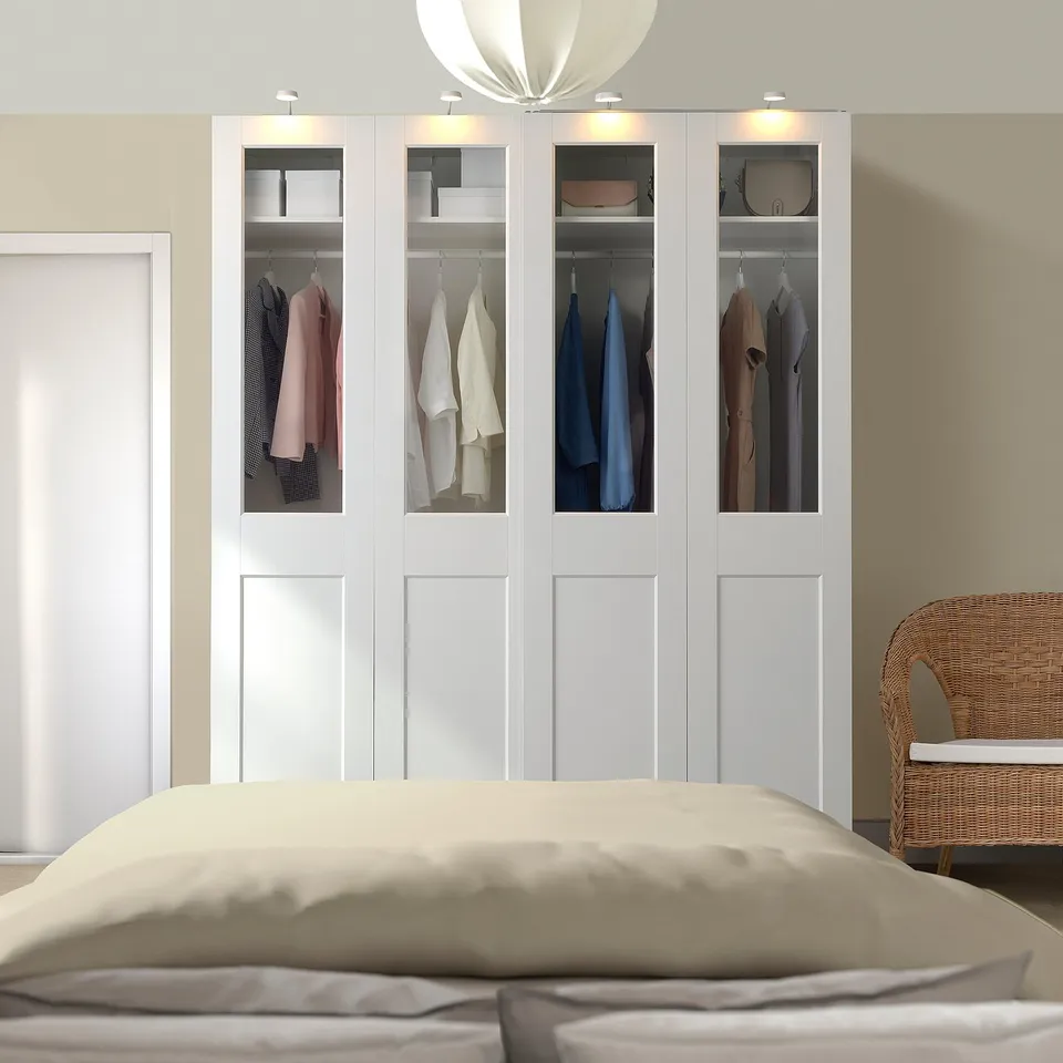 White PAX wardrobe with glass doors, displaying clothes. Tall, modern, and efficient, next to bed and wicker chair.