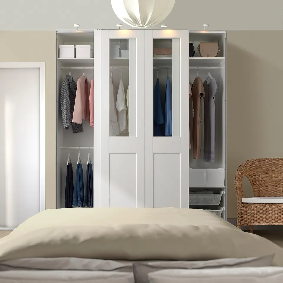 A white PAX wardrobe with glass doors and inside lights. Its large, tall, and wider than a bed, holding clothes and accessories neatly.
