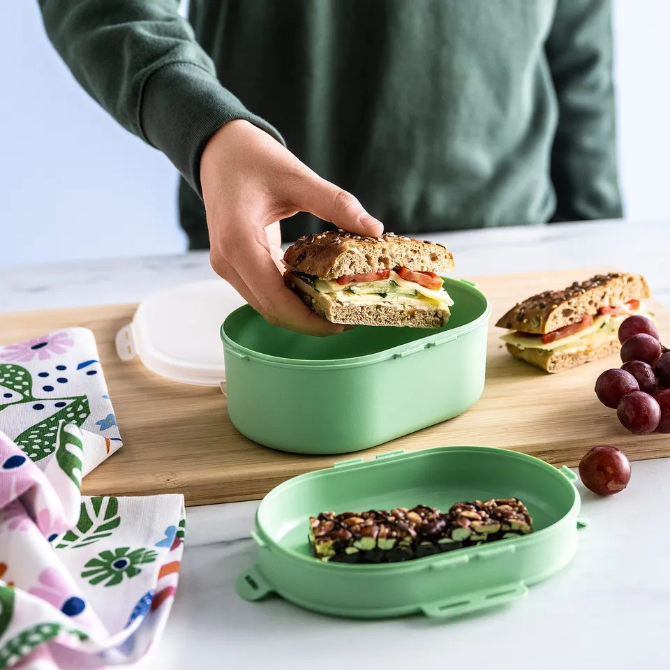 A person puts a sandwich in a 2-tier green lunch box on a table with snacks and grapes, next to a patterned cloth.