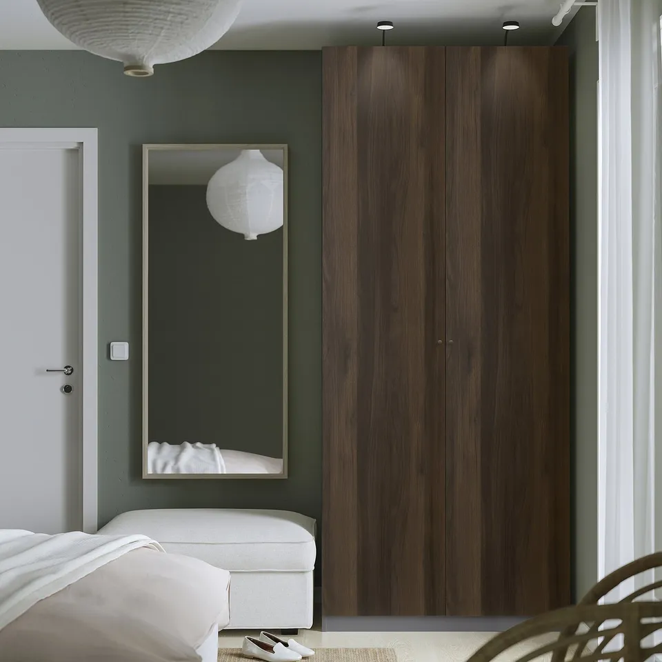 Modern bedroom with tall dark wood PAX wardrobe, sage green wall, large mirror, white bed.