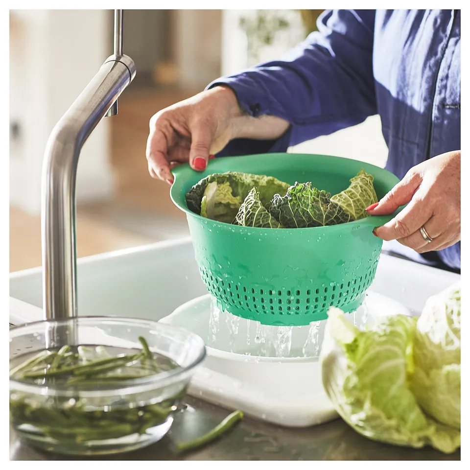 A person rinses green vegetables in a green UPPFYLLD colander under a kitchen tap.