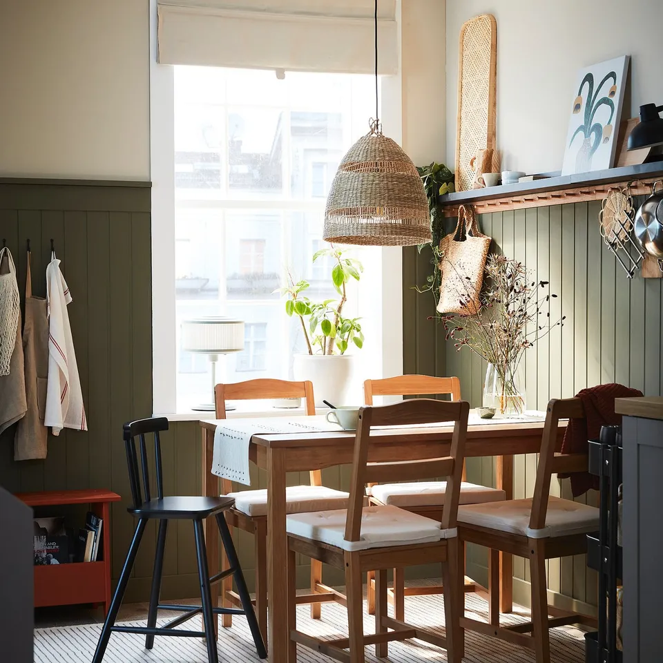 Bright kitchen with wooden table, chairs, and woven pendant lamp. Large window lets in natural light. Plants and decorative items on the windowsill.