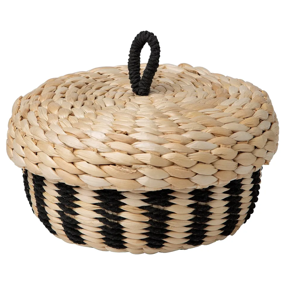 An IKEA MÄVINN woven basket with lid. Its beige with black accents, round, and handcrafted from banana fibres.