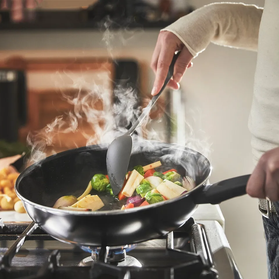 Person cooking vegetables in a non-stick frying pan on a stove, steam rising.