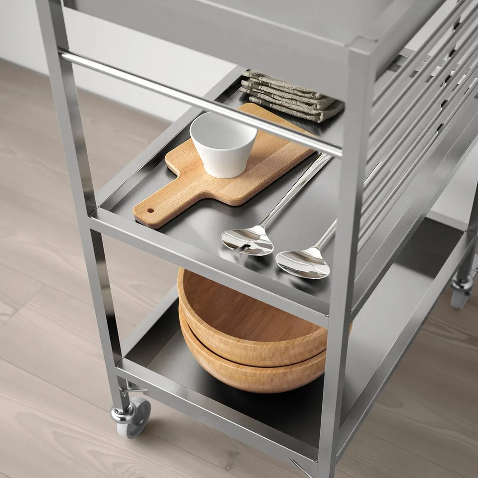 Modern metal kitchen trolley with wheels, holding wooden bowls, spoons, cup, and cutting board.