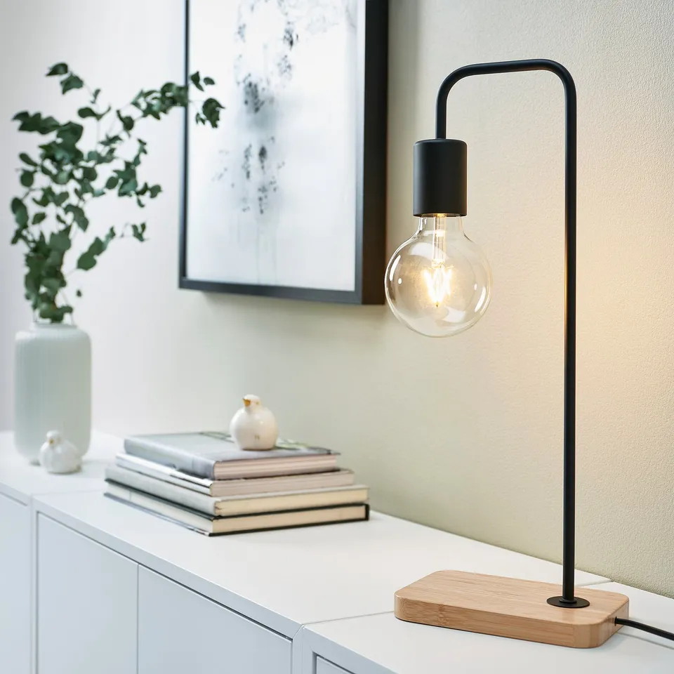 Black TVÄRHAND lamp on table with books and plants, minimalist and tall.