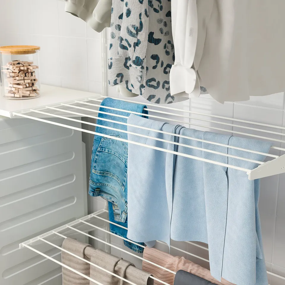 A white BOAXEL drying rack holds light blue jeans and a beige garment, organised and air-drying in a laundry room.