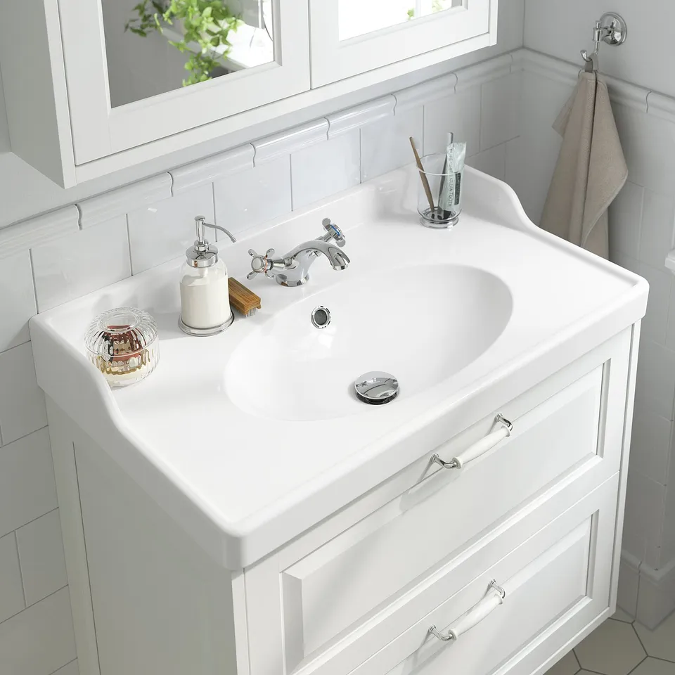 White bathroom sink with chrome-plated tap, soap dispenser, and toothbrush holder, set against white tiled walls and mirror.
