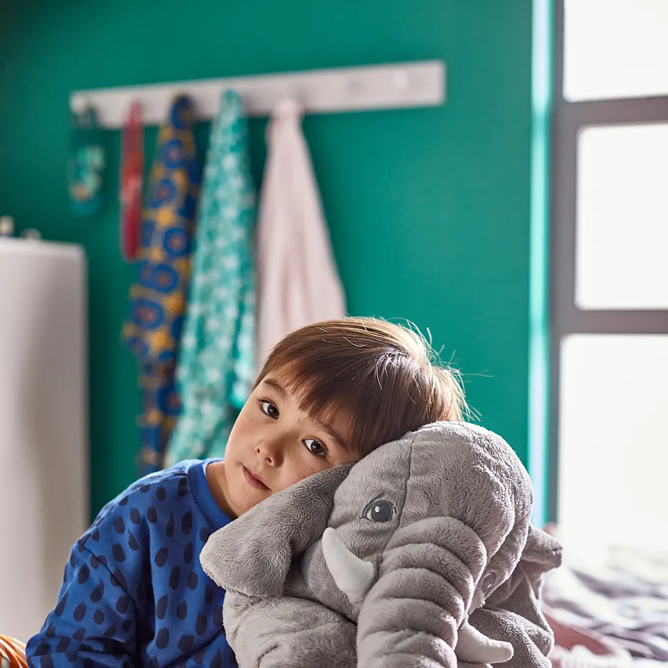 Child sitting on bed hugging large grey JÄTTESTOR elephant cuddly toy. Blue polka dot pyjamas, striped socks. Cosy, green-walled room.