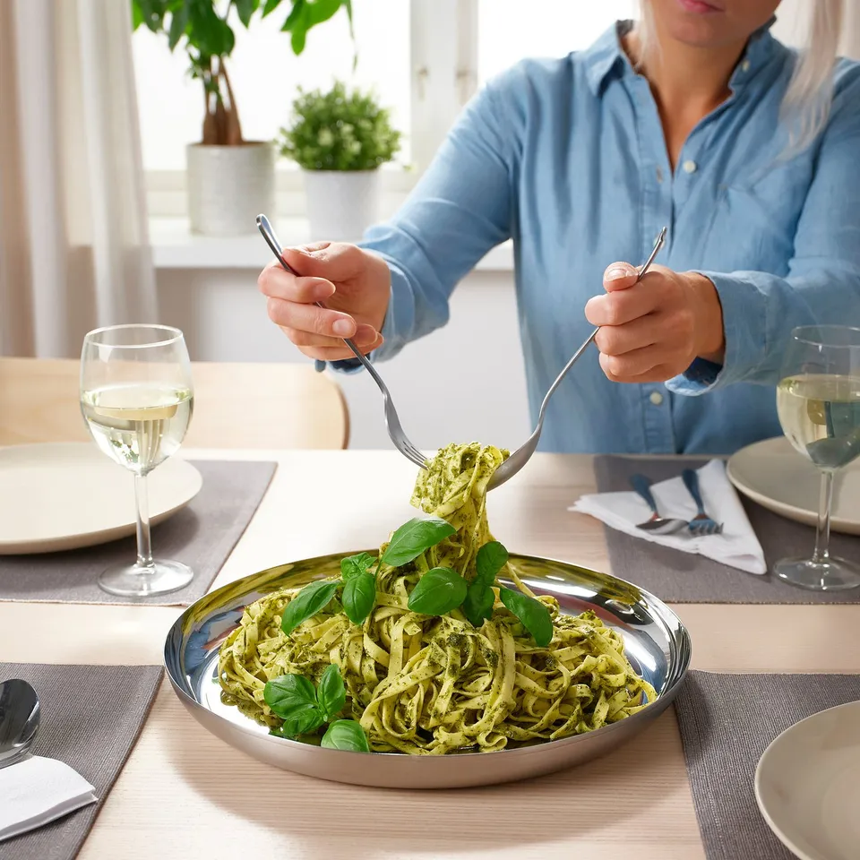 Person twirling pasta from a large stainless steel serving plate, with glasses of wine and dining table setup.
