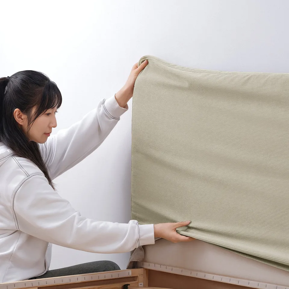 A person is putting on a light beige bed frame cover with a hanging skirt, showing its easy-to-remove feature.