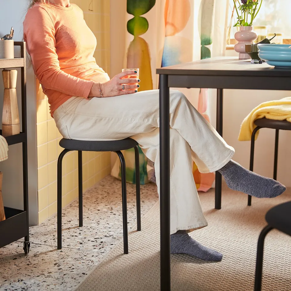 Person sits on GULTARP stool, holding drink, showcasing its comfort and space-saving design.