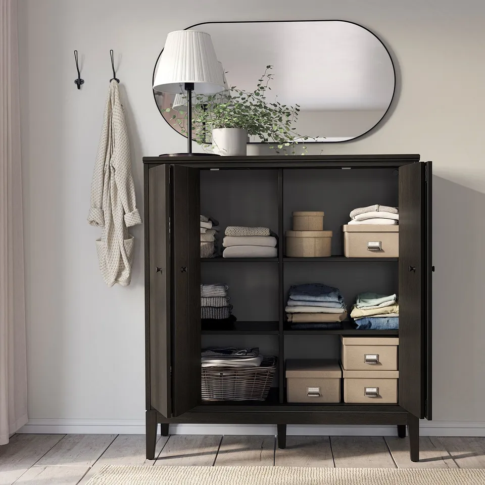 Black IDANÄS cabinet with clear doors, mirror, lamp, plant, clothes & boxes, light wall, towel nearby.
