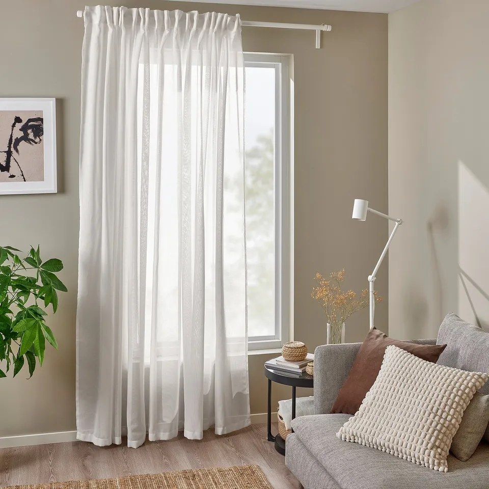 Bright room with white ÄNGSFRYLE sheer curtains, wide & airy, letting light through. Accented with cosy cushions and a modern lamp.