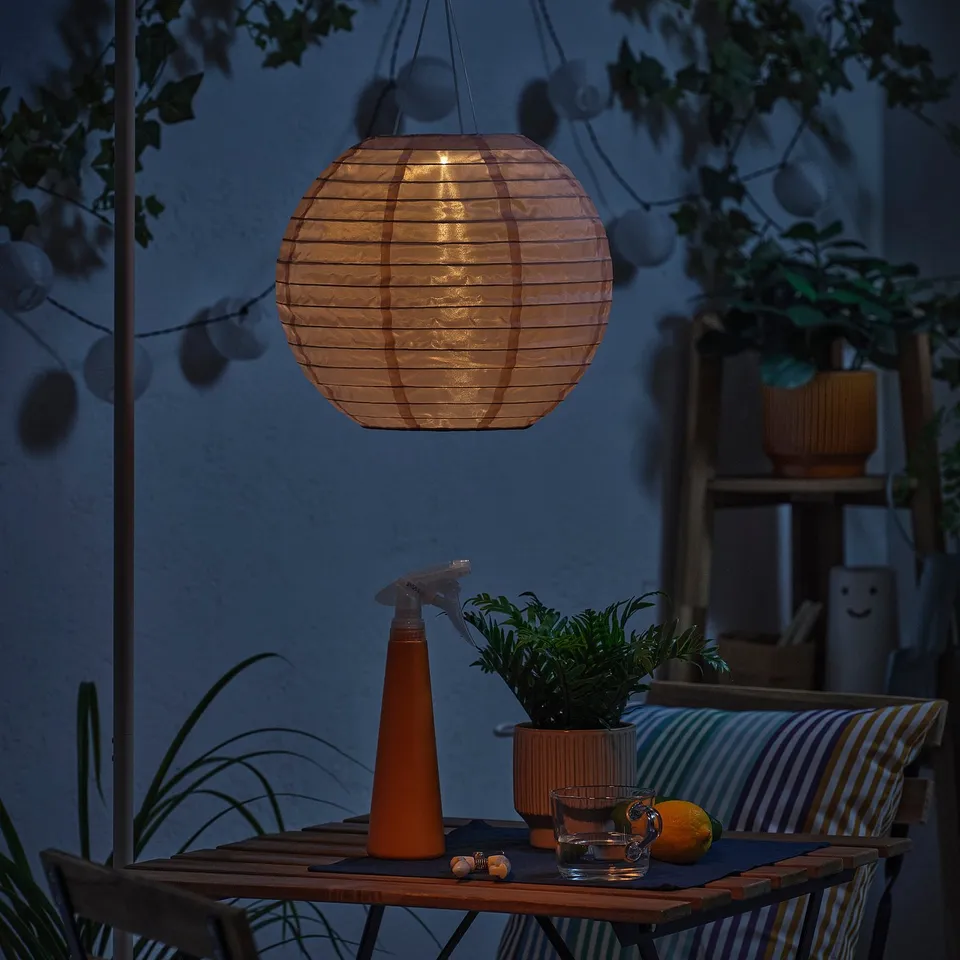 Cosy outdoor setting with a SOLVINDEN solar-powered lantern casting warm light over a table with plants, drinks, and an orange.