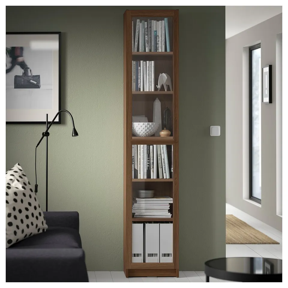 Tall wooden bookcase with multiple shelves, holding various books and decorative items, next to a black and white patterned chair.