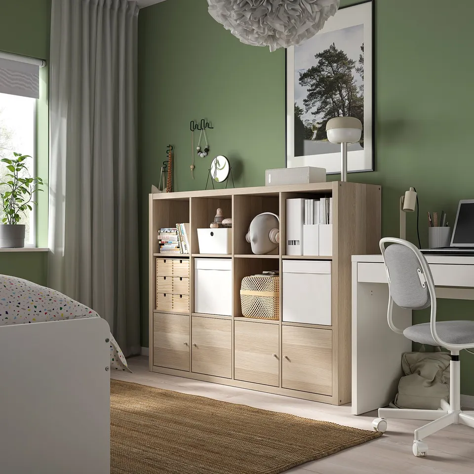 Modern bedroom with KALLAX shelves, green walls, multifunctional storage, white desk and grey chair, natural light from a window.