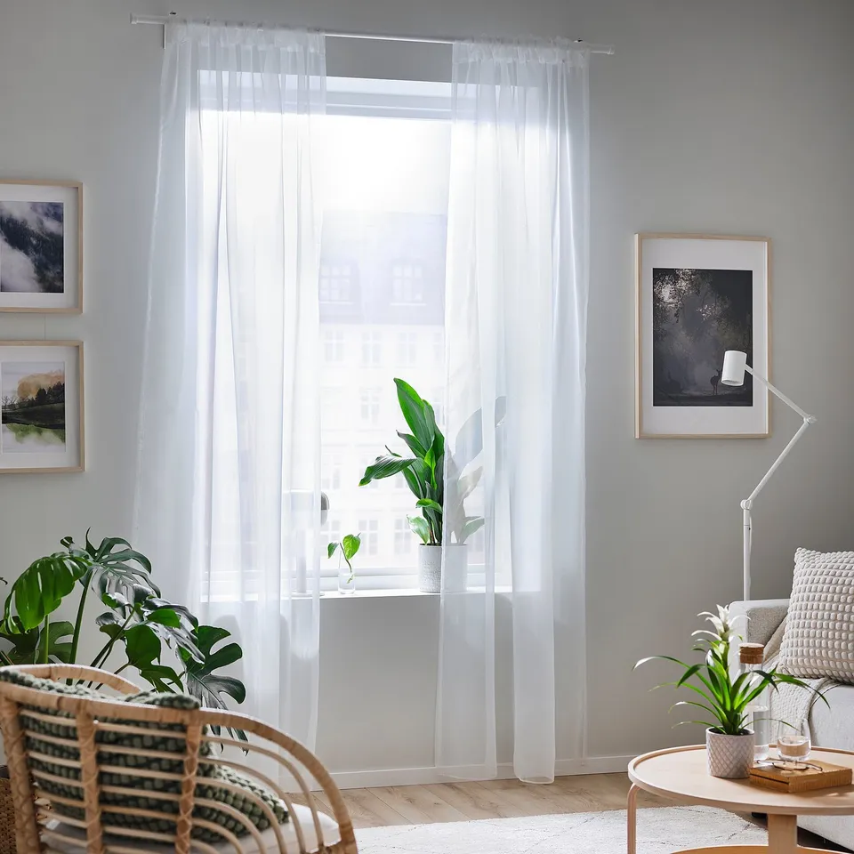 Bright room with white curtains, wicker chair, plant, table, books, art frames.