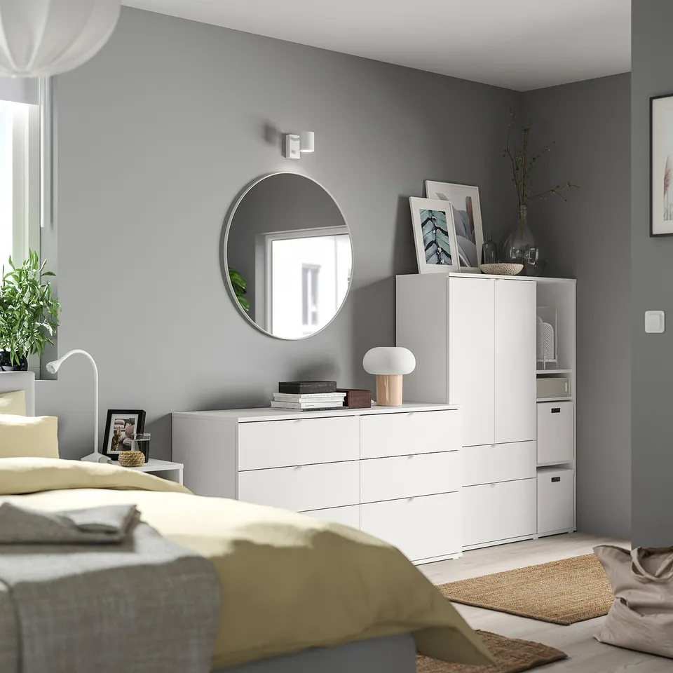 Modern bedroom with grey walls, white furniture, yellow blanket, round mirror, lamp, and decorative items.
