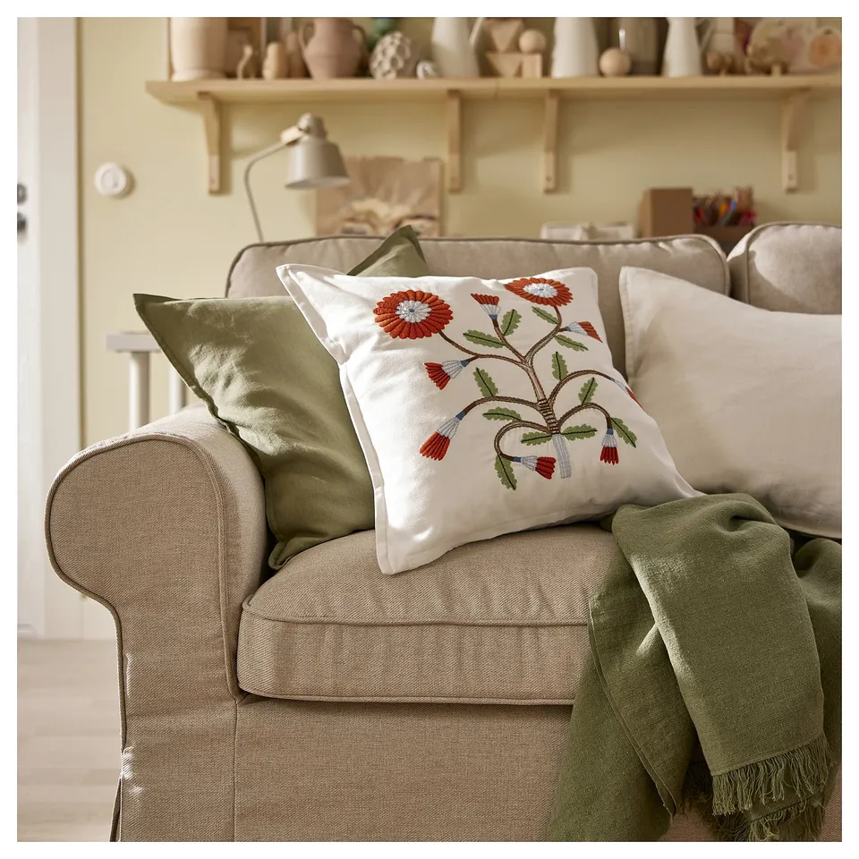 Living room with beige sofa, colourful pillows, green blanket, and floral pillow FACELIOR. Shelves with neutral decor in background.