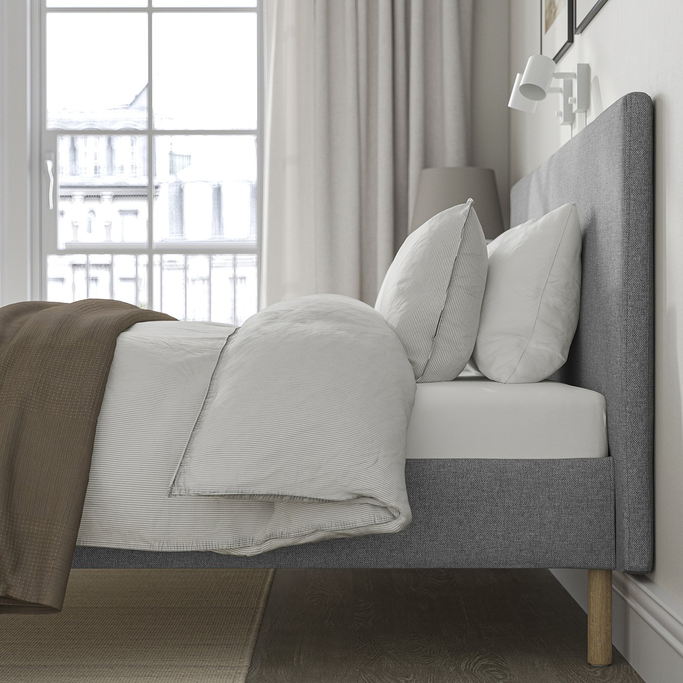 Bed near window with grey headboard, white pillows, folded blanket, wooden legs, and wall light.