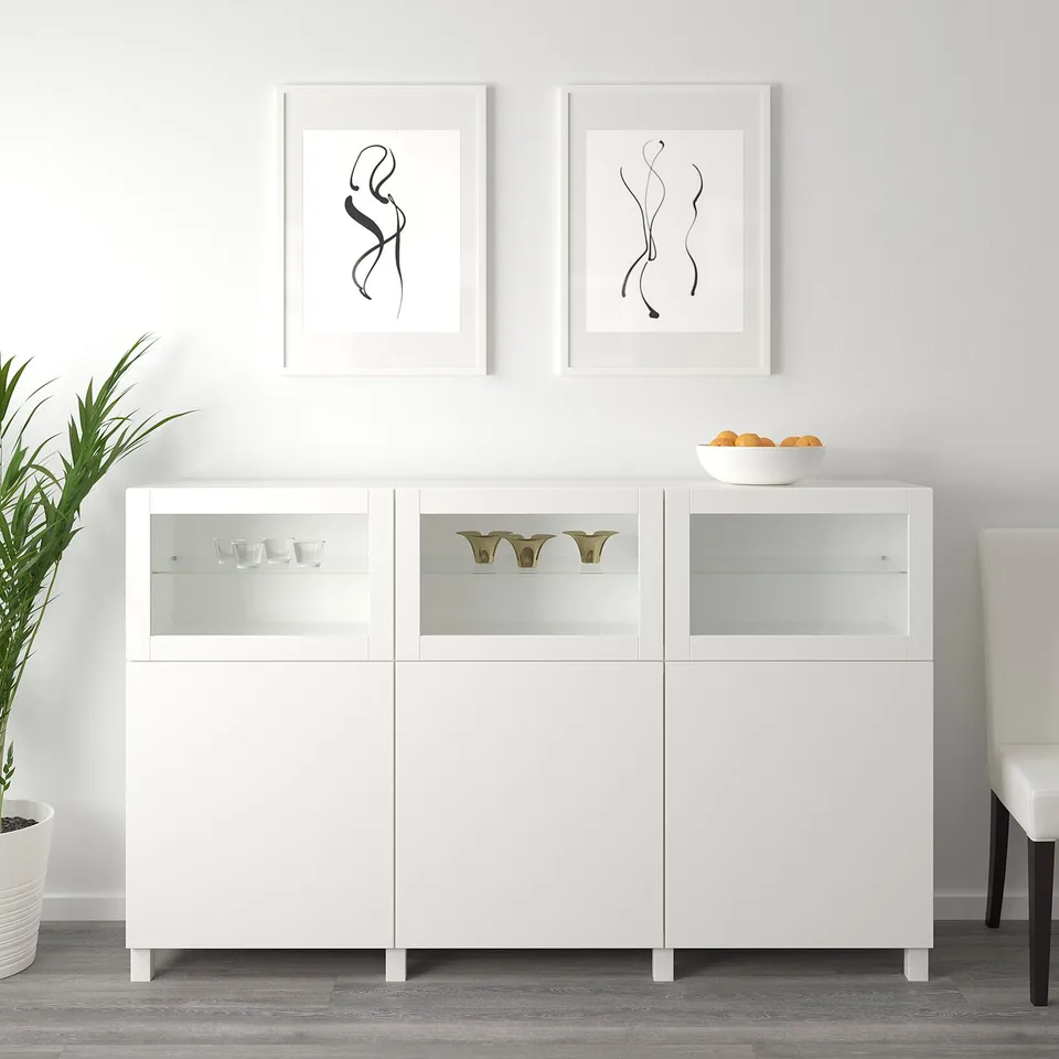 A white BESTÅ sideboard with glass doors, displaying items inside, and a bowl on top, set against a wall with abstract art.