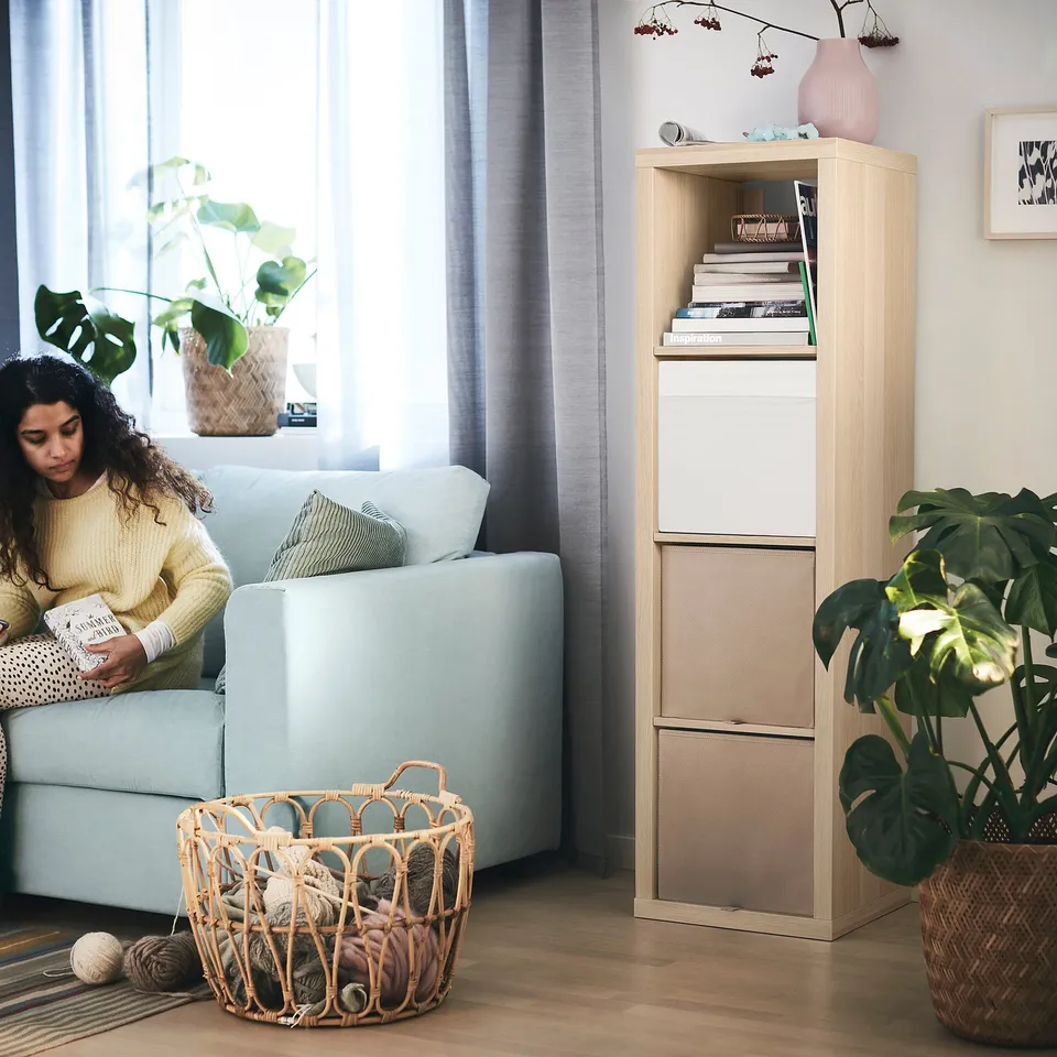 Cosy living room scene with person reading on light blue couch, beside woven basket of yarn, near KALLAX shelving unit with books and decor.