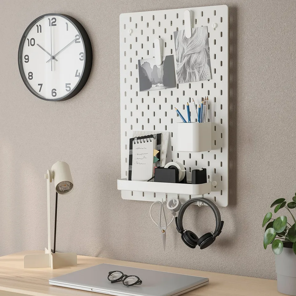 Wall-mounted pegboard with accessories including containers, photos, headphones, and clock. Organises office supplies and personal items efficiently.