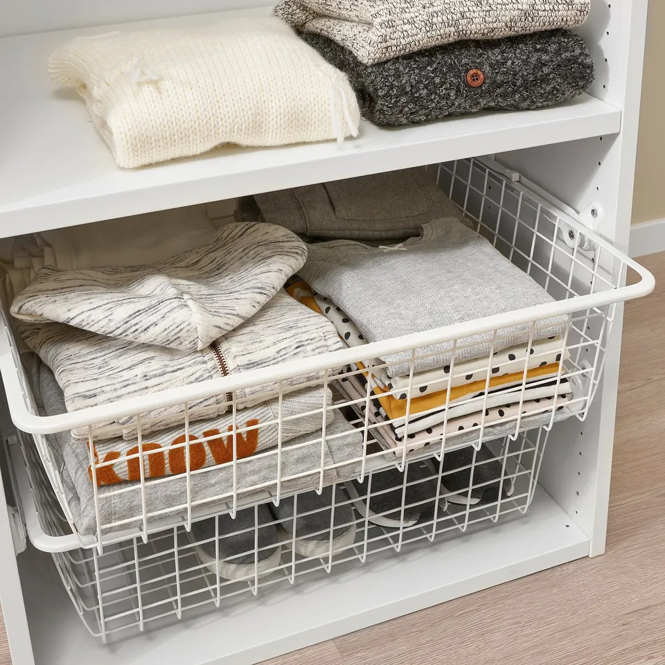 White storage shelf with wire basket holding neatly folded clothes, part of IKEAs HJÄLPA organisational system.