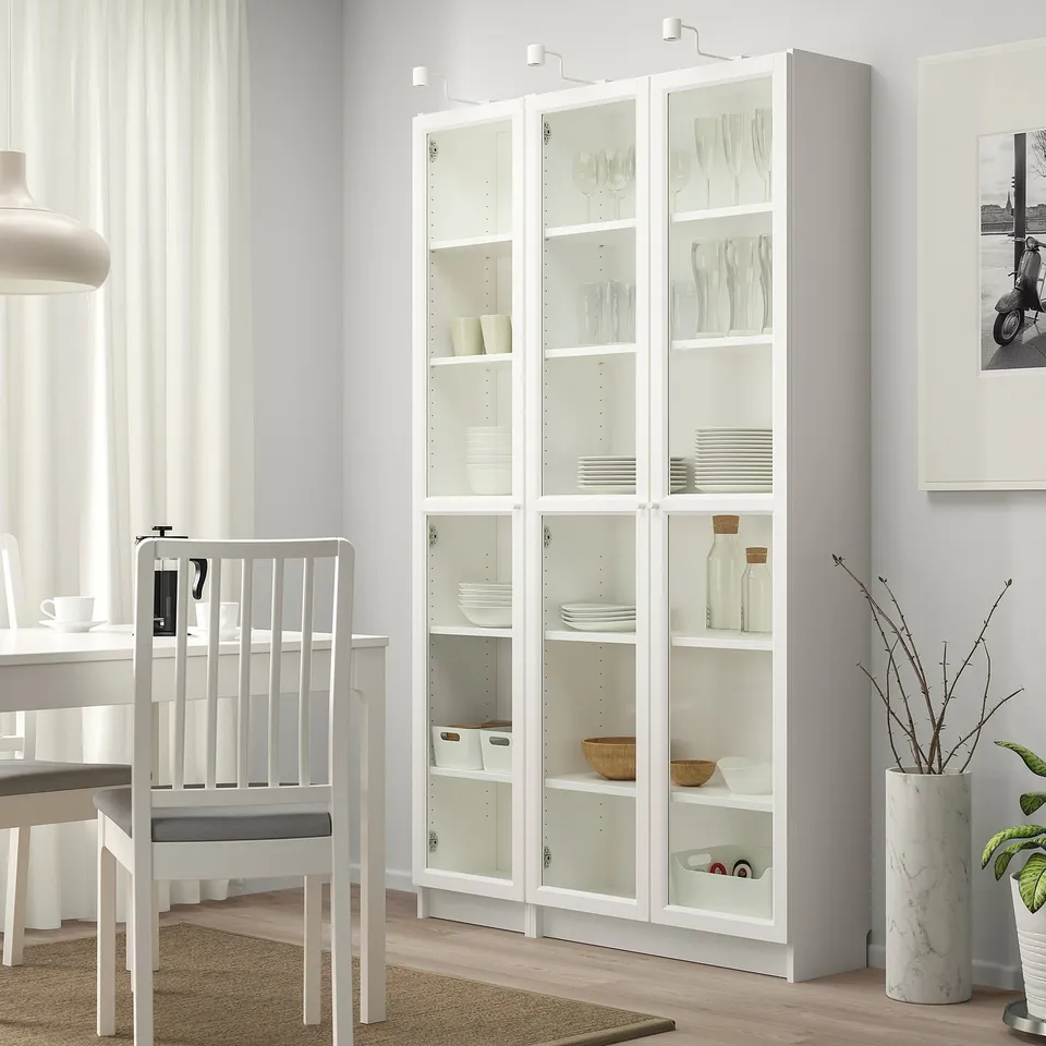 White BILLY bookcase with glass doors, near dining table and chair, holding dishes and decor, almost reaching ceiling.