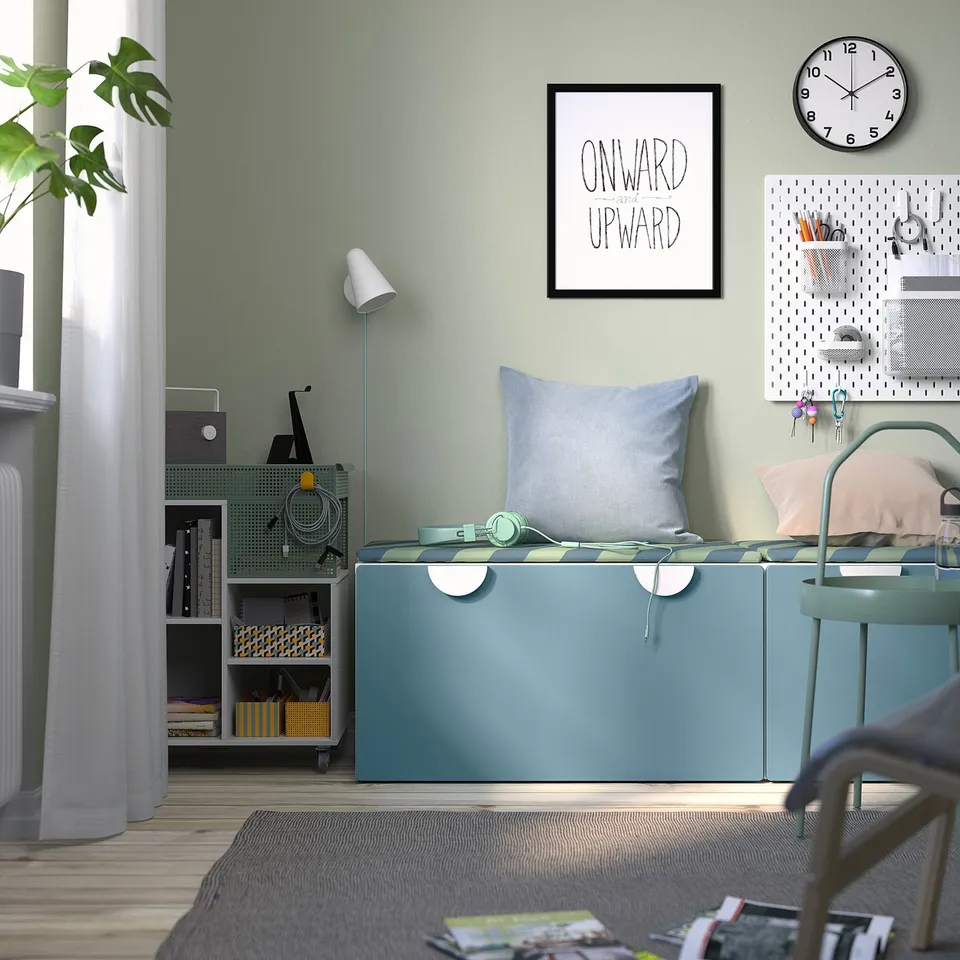 Small blue storage bench under framed poster, near organised shelving, with headphones, & wall clock.