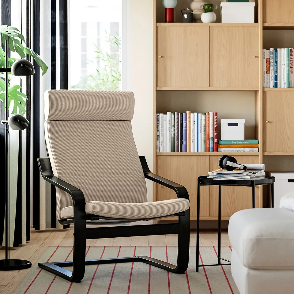 Modern living room with poang armchair, black frame, beige cushion, wooden bookcase filled with books and decor, striped rug, and floor lamp.