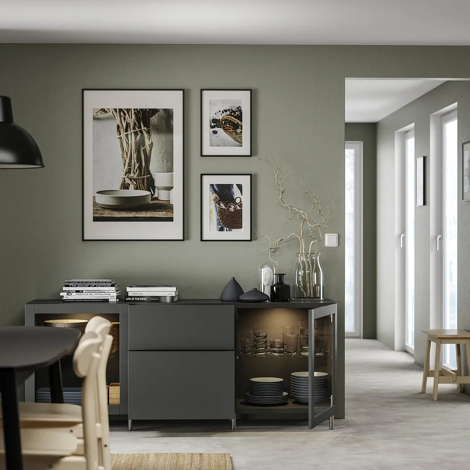 Modern dining room with BESTÅ storage in black holding dishes and vases, against sage green wall adorned with framed photos and a light fixture.
