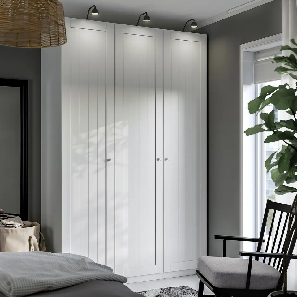 Modern bedroom with tall white PAX wardrobe, lights, rug, chair, bed, linens basket, and large plant.