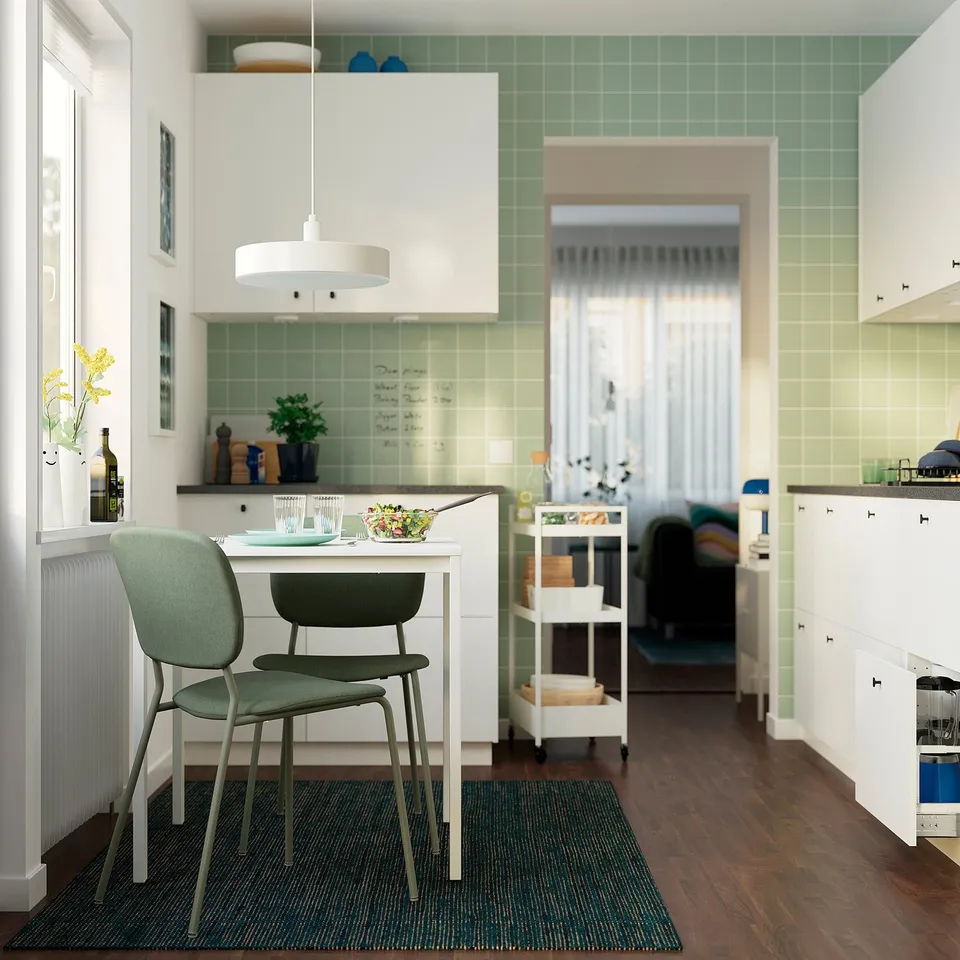 Modern kitchen with green tiles, white cabinets, and a compact dining set with salad, glasses, and a wine bottle on the table.